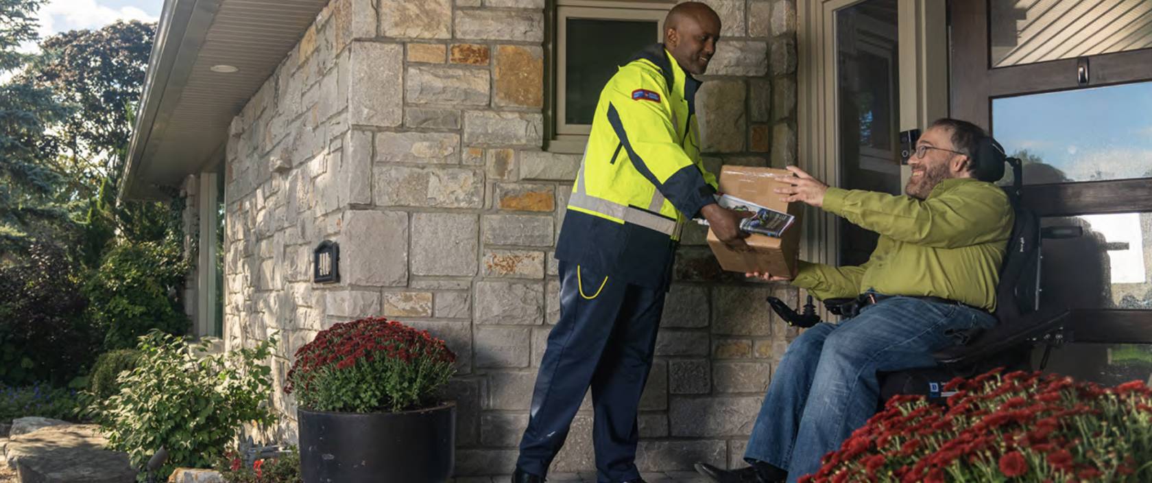 A Canada Post delivery agent hands a package to a man in a wheelchair at his front door. 