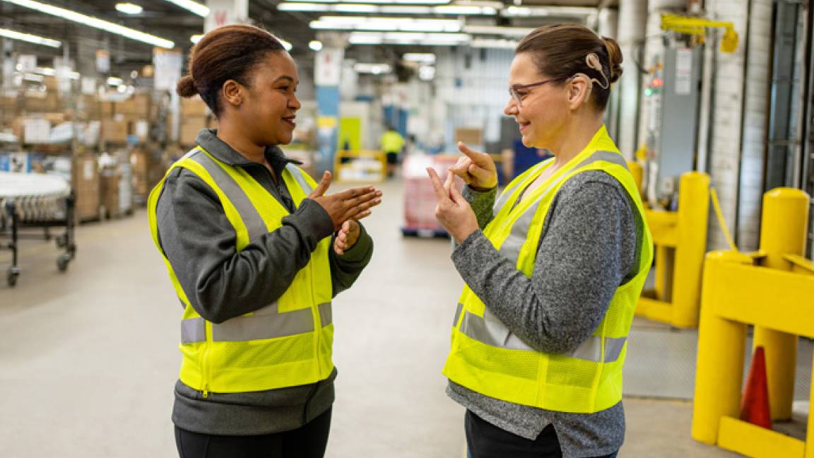 Two employees in a Canada Post sorting facility communicate in sign language, one has a visible hearing aid.
