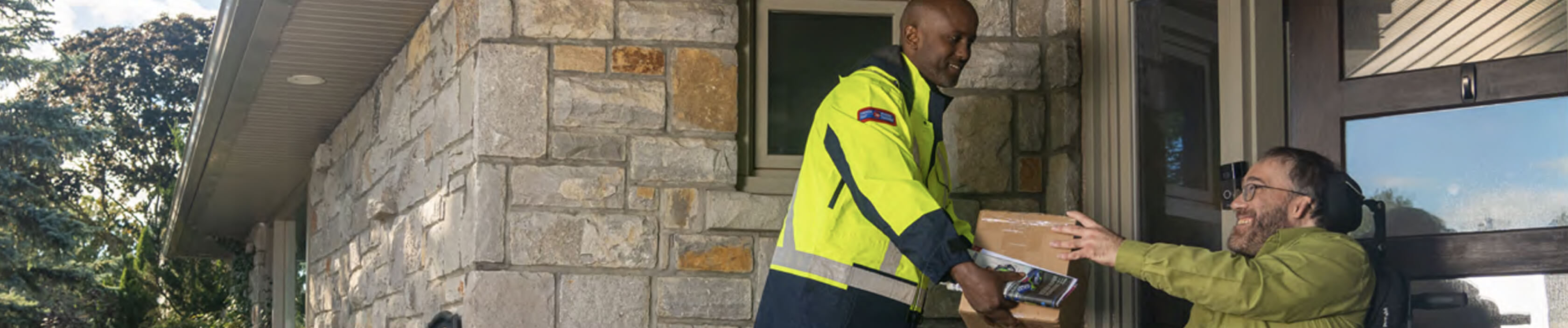 A Canada Post delivery agent hands a package to a man in a wheelchair at his front door. 