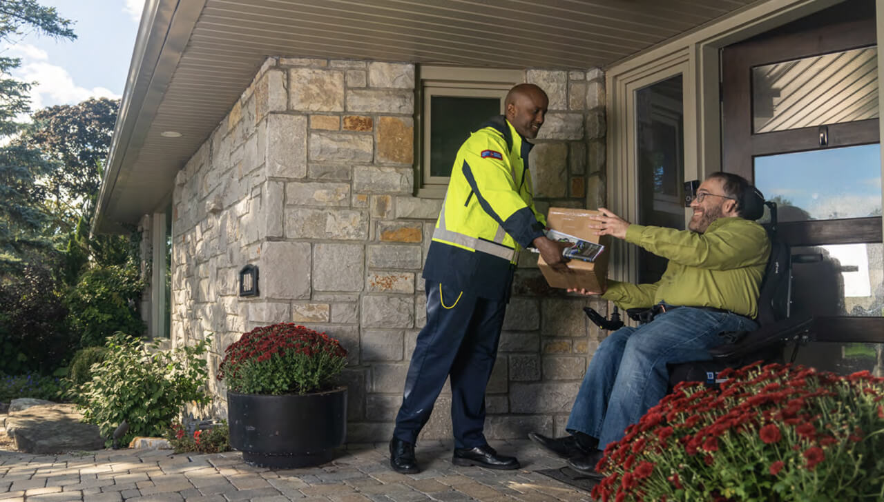 A Canada Post delivery agent hands a package to a man in a wheelchair at his front door. 