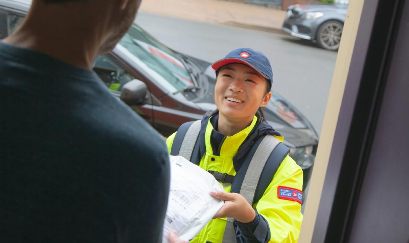 A Canada Post employee wearing a bright yellow jacket delivers a package to a man standing in his doorway. 