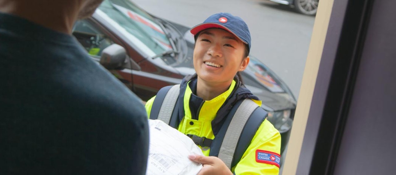 A Canada Post employee wearing a bright yellow jacket delivers a package to a man standing in his doorway. 