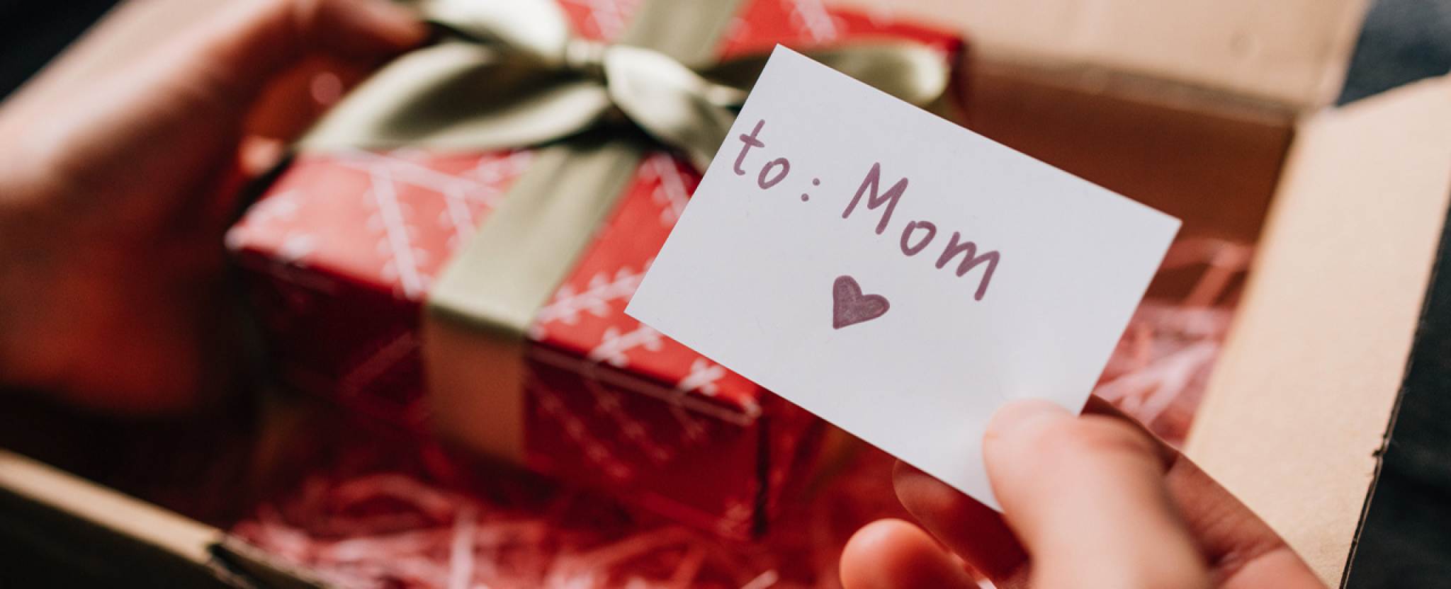 Hands open a shipping box which contains a wrapped gift box and a card that reads 'to Mom.’