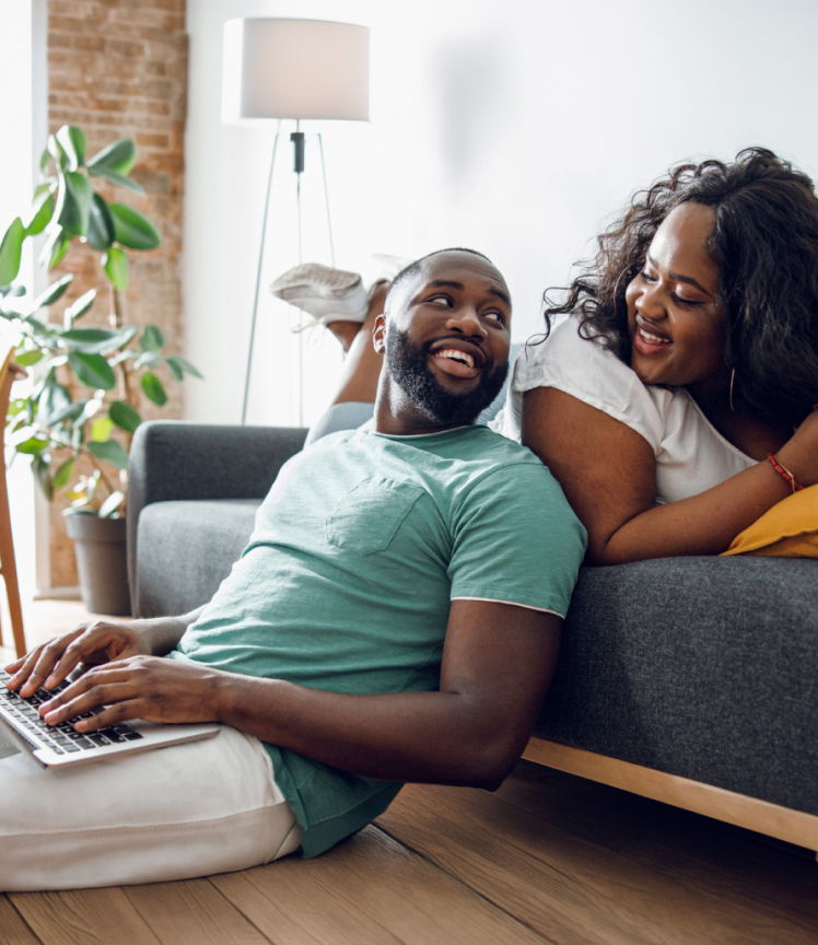 A man types on a laptop and leans against a couch a woman is sitting on. They smile at each other.