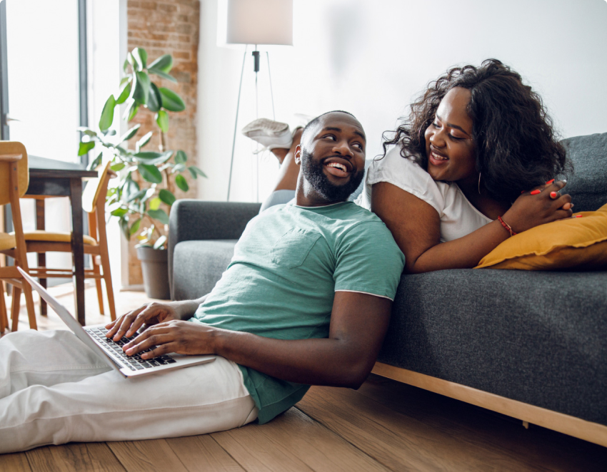 A man types on a laptop and leans against a couch a woman is sitting on. They smile at each other.