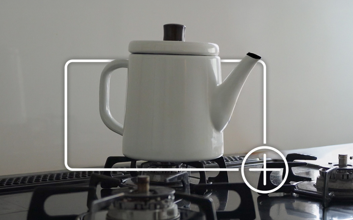 White enamel kettle on a gas stove burner framed by a white focus box with a plus icon indicating adding the item to an online shopping cart.