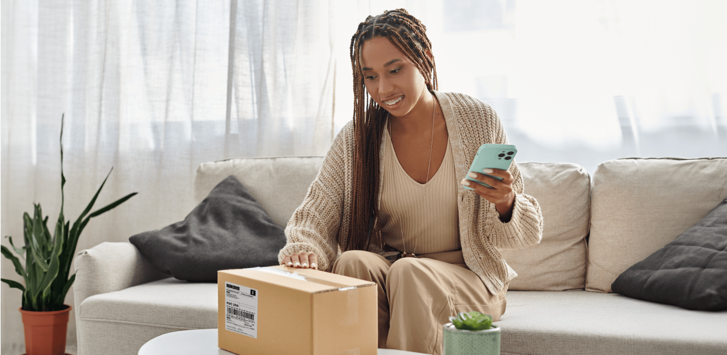 A smiling woman looks at her smart phone as she holds a package.