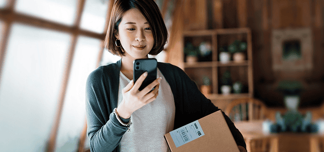 A woman looks at her mobile phone and smiles, holding a package with a return label under her arm.
