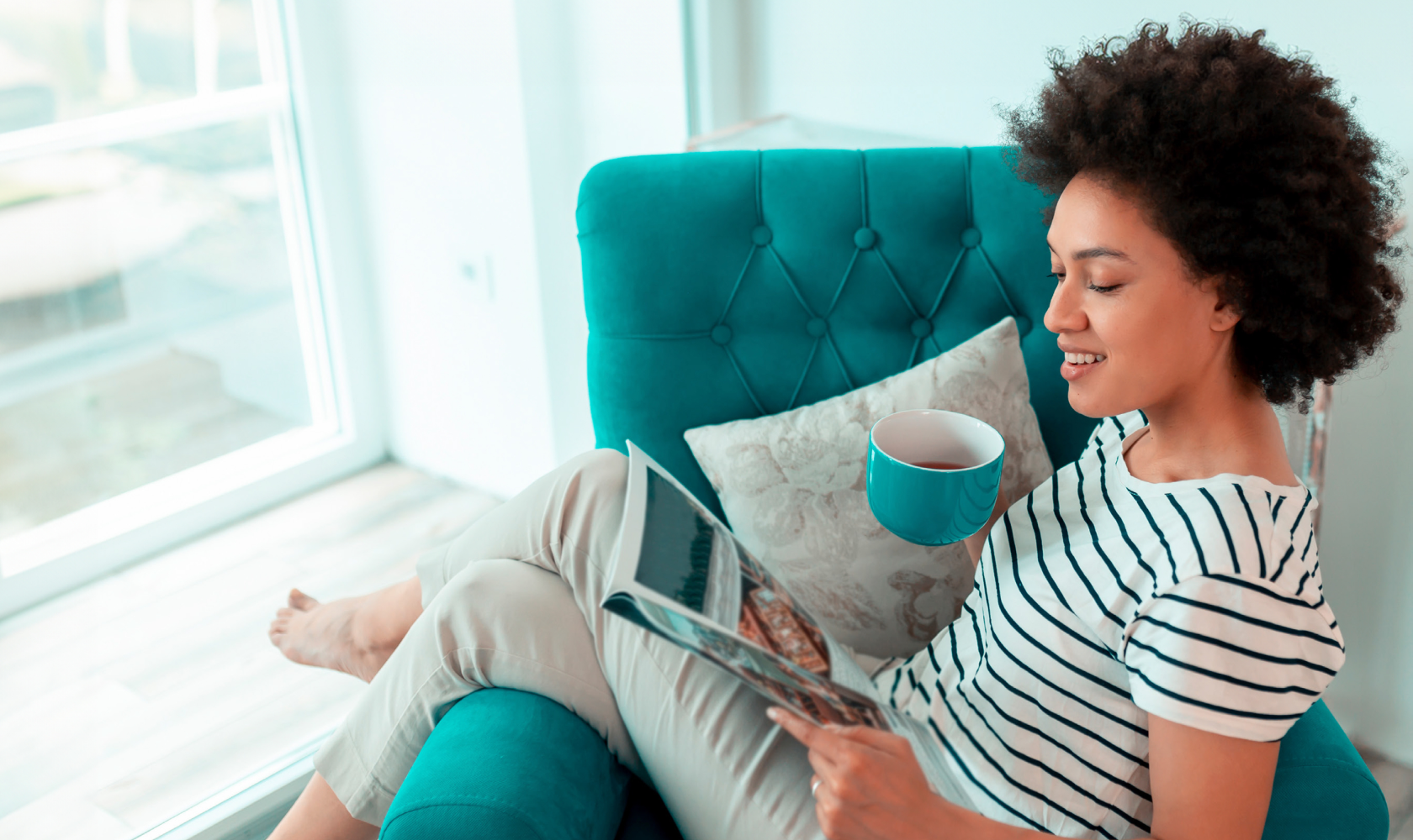 A woman sits in a chair reading a catalogue. She holds a teal mug of tea that matches the chair she is sitting in.