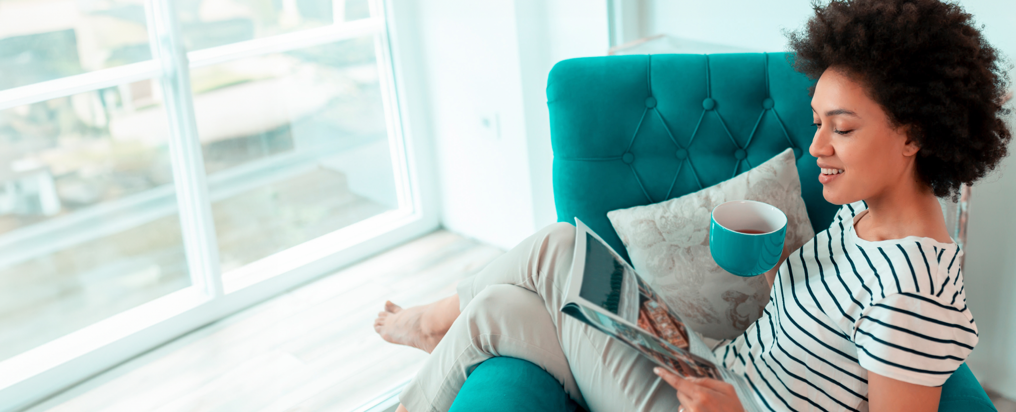 A woman sits in a chair reading a catalogue. She holds a teal mug of tea that matches the chair she is sitting in.