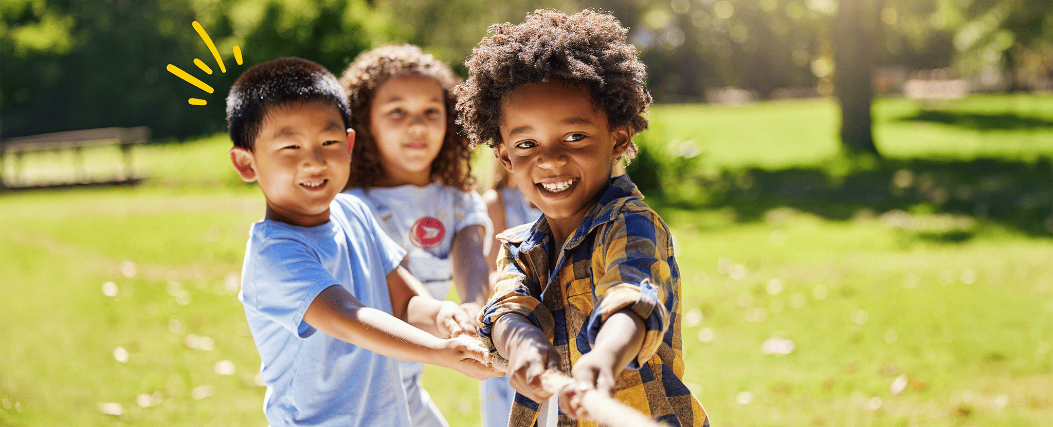 Children playing tug-of-war outdoors, pulling a rope together on a sunny day with grass and trees in the background.