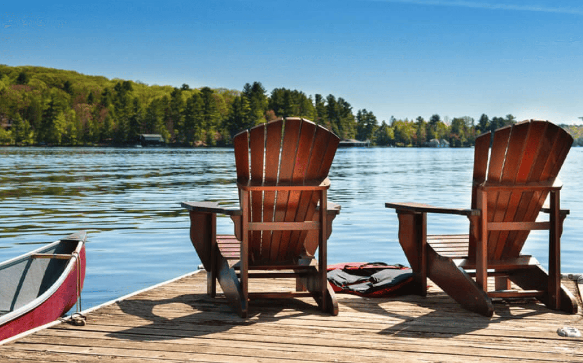 Two wooden chairs sit on a lakeside dock, next to a canoe.