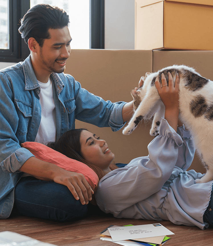 A smiling couple play with a cat on the floor. There is a small stack of mail beside them and stacked cardboard boxes in the background.