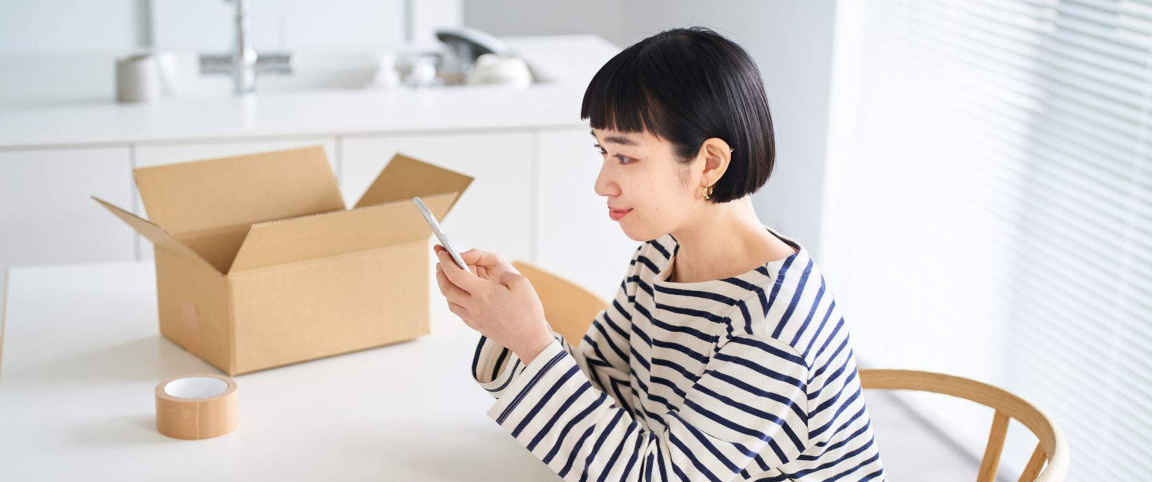 A woman types on her mobile phone. She sits at a table that has an open shipping box and a roll of tape on top.