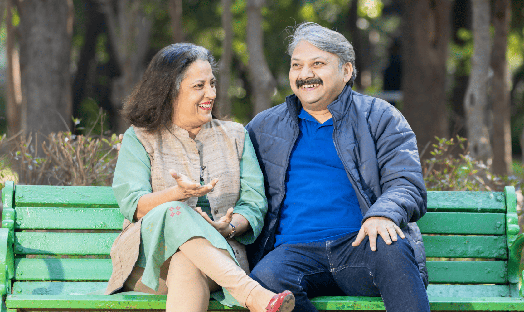 A mature couple smile as they sit on a park bench together. 