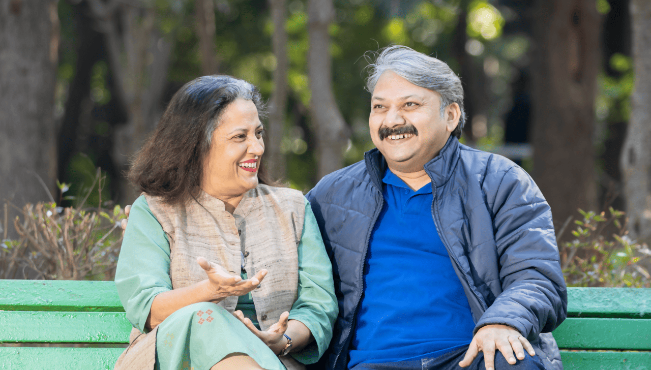 A mature couple smile as they sit on a park bench together. 