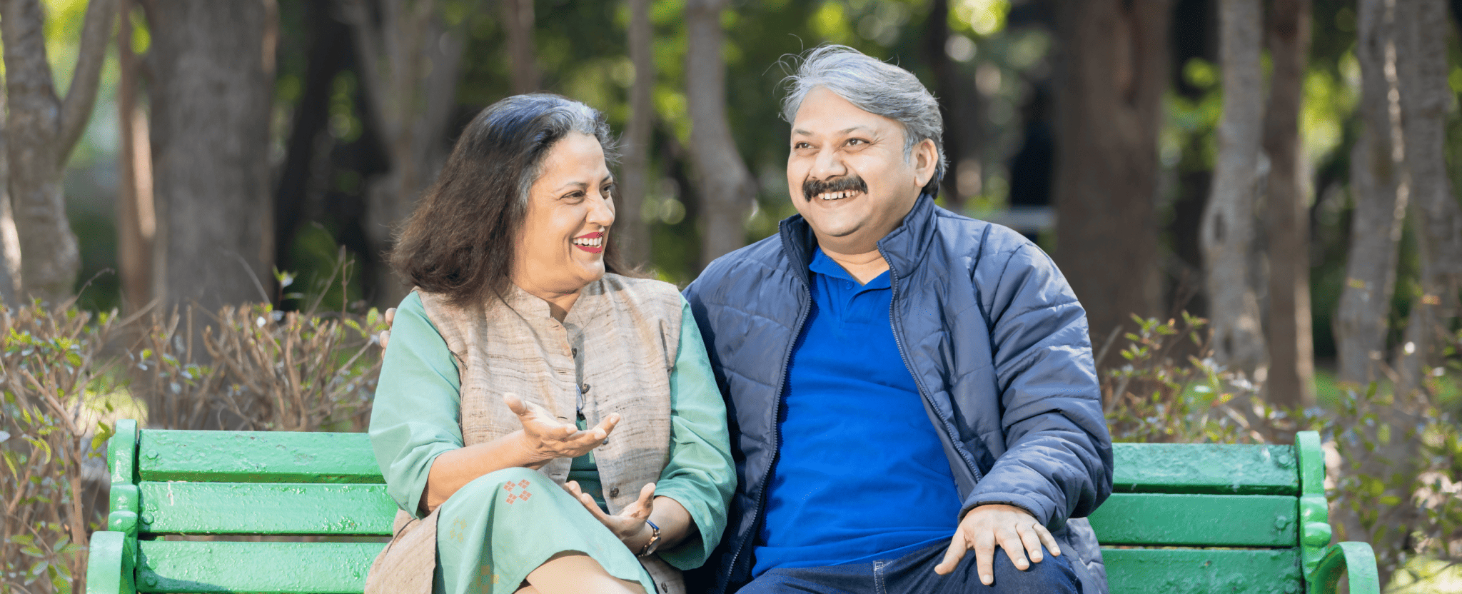 A mature couple smile as they sit on a park bench together. 
