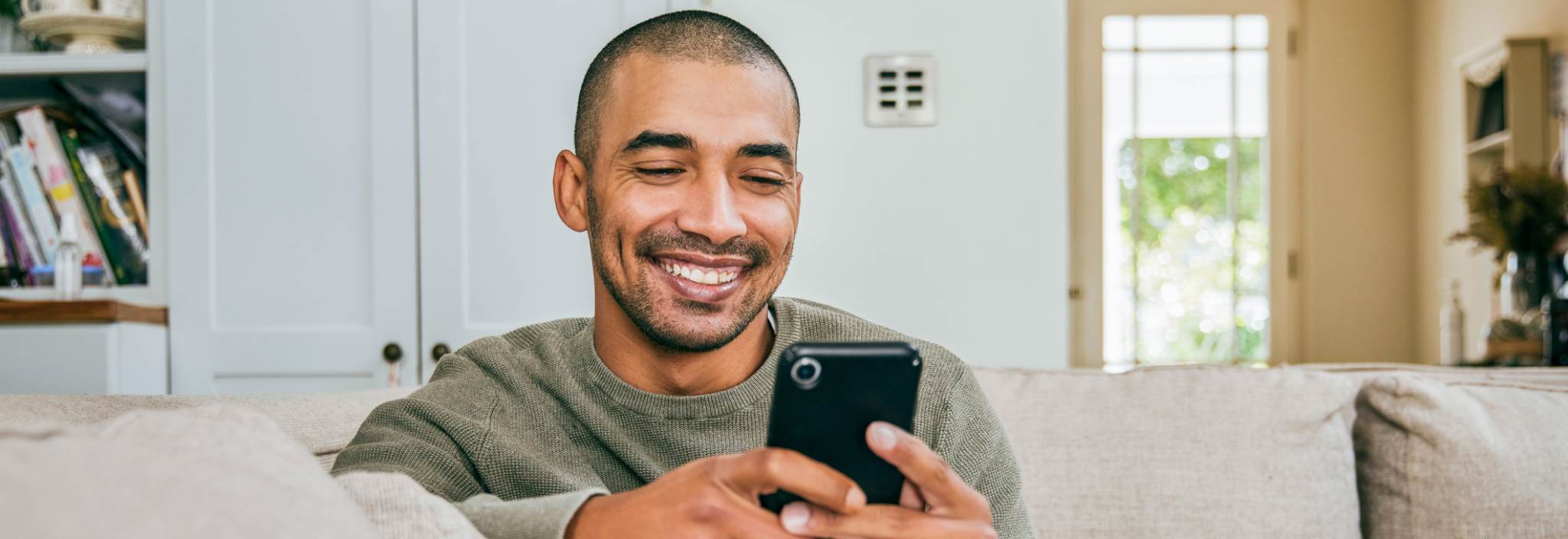 A young man smiles and stares a cellphone in his hands. He sits on a couch in his living room.