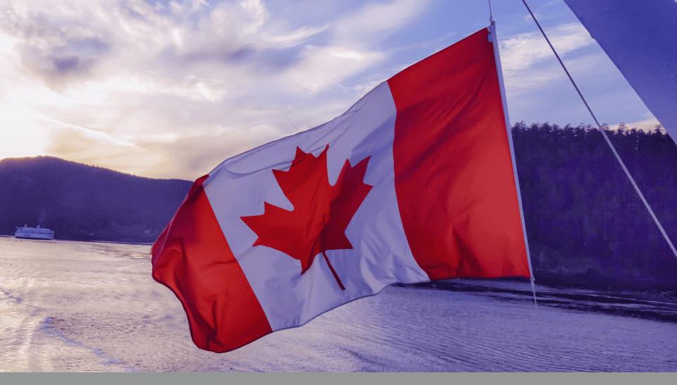 A Canadian flag waving with a body of water and forested hills in the background at sunset.