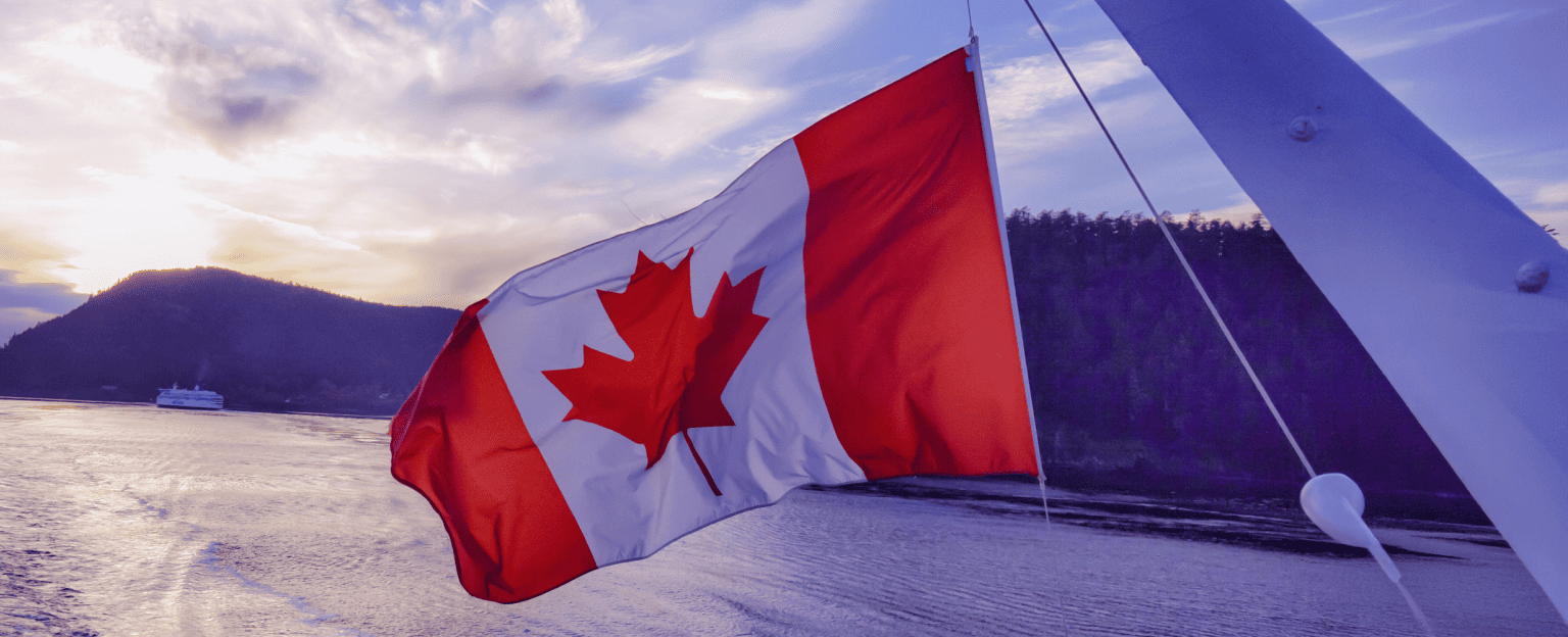 A Canadian flag waving with a body of water and forested hills in the background at sunset.