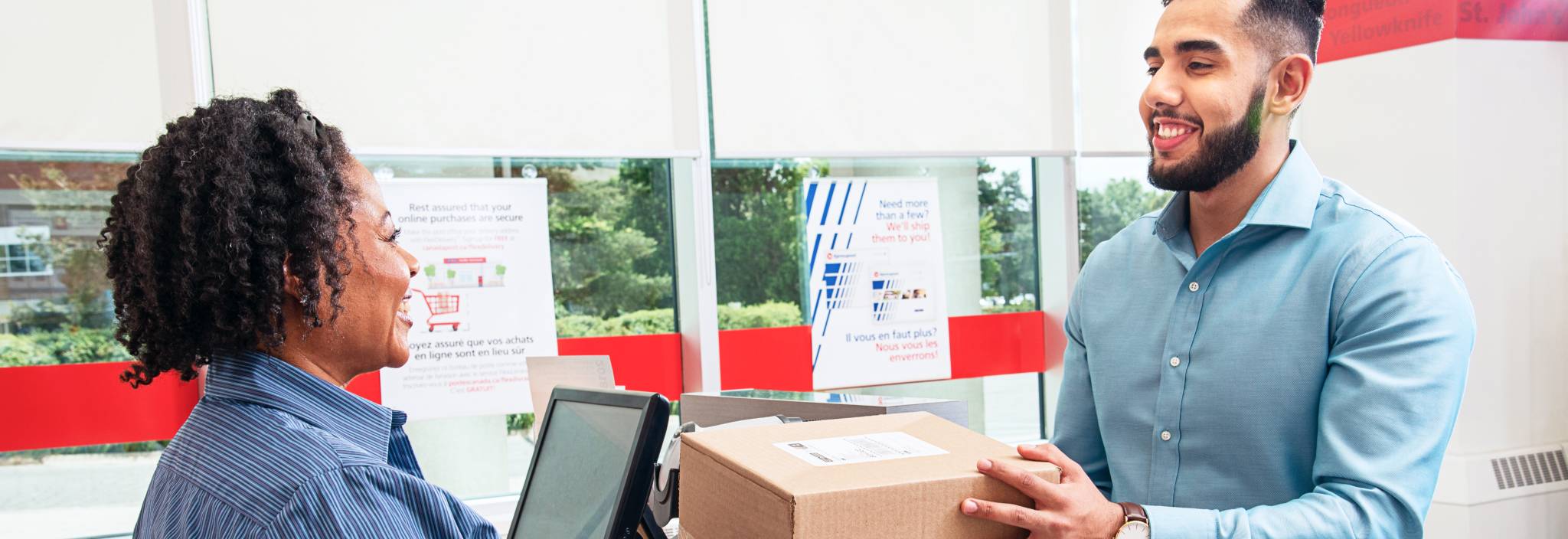 A smiling Canada Post retail employee hands a package to a man from behind a post office counter.