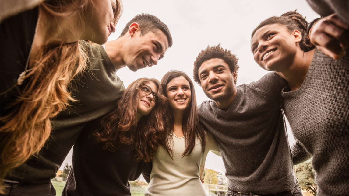 A diverse group of young adults smile and huddle together with their arms around each other's shoulders.