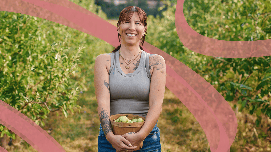 Une femme debout dans un champ tient un panier de fruits en souriant.