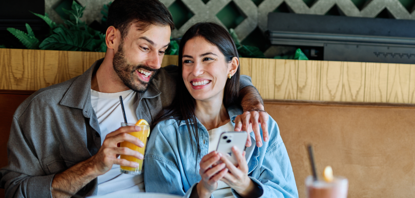 Un couple est assis dans un café avec des plantes en arrière-plan; la femme tient un téléphone et l’homme une boisson.