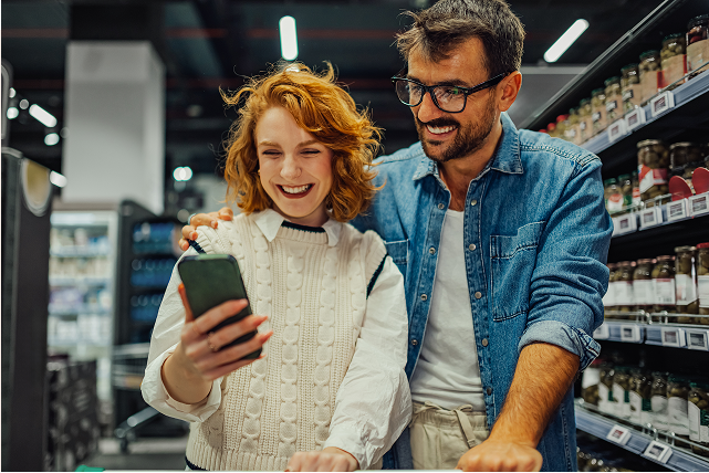 Une femme et un homme derrière un panier d'épicerie consultent un téléphone intelligent, avec des étagères en arrière-plan.