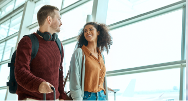 Un couple avec des valises discute en marchant dans un terminal d’aéroport alors que la lumière du soleil passe à travers de grandes fenêtres et qu’un avion est visible à l’extérieur.