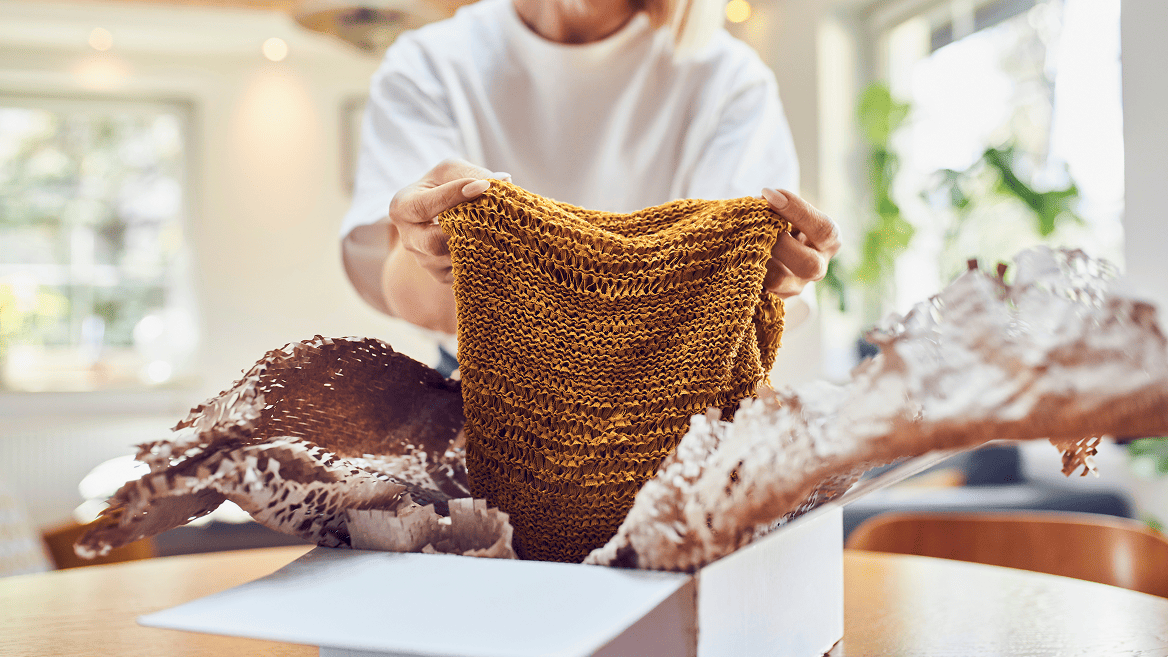 Person unpacking a knitted mustard coloured garment from an open box with packing paper on a table. 