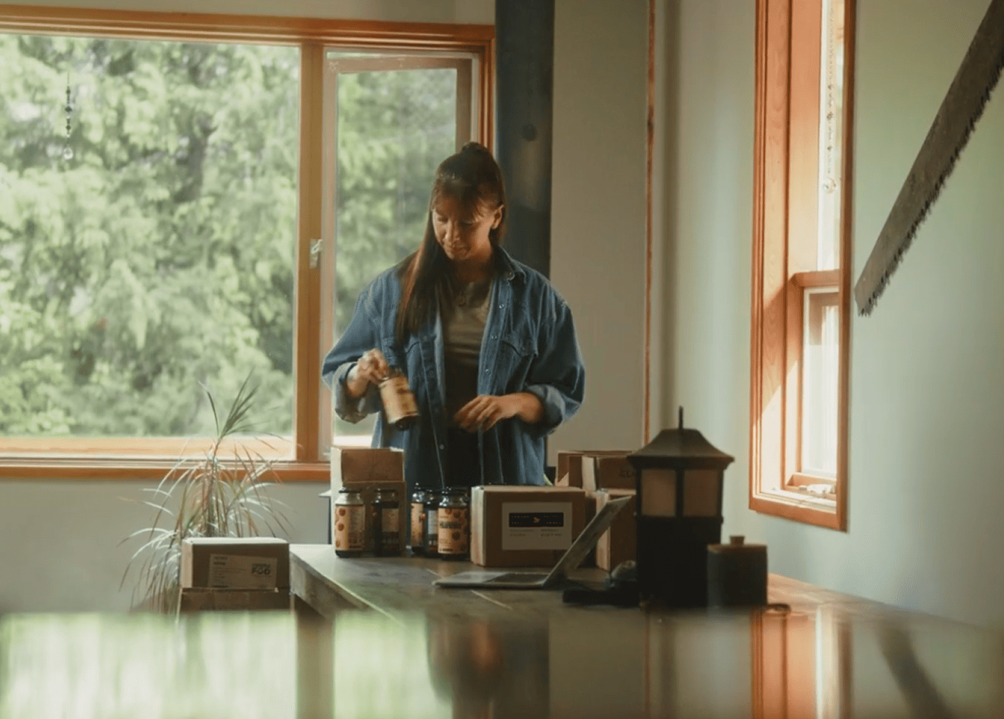 Venessa Stonehouse, Co-Founder and CEO of Mindful Food, places labeled jars into shipping boxes on a table beside a laptop in a sunlit workspace.