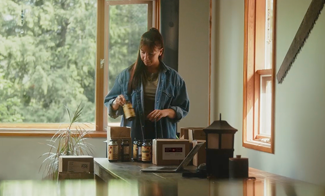 Venessa Stonehouse, Co-Founder and CEO of Mindful Food, places labeled jars into shipping boxes on a table beside a laptop in a sunlit workspace.