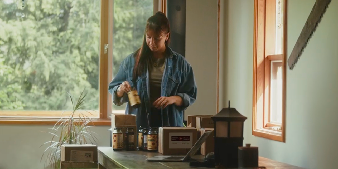 Venessa Stonehouse, Co-Founder and CEO of Mindful Food, places labeled jars into shipping boxes on a table beside a laptop in a sunlit workspace.