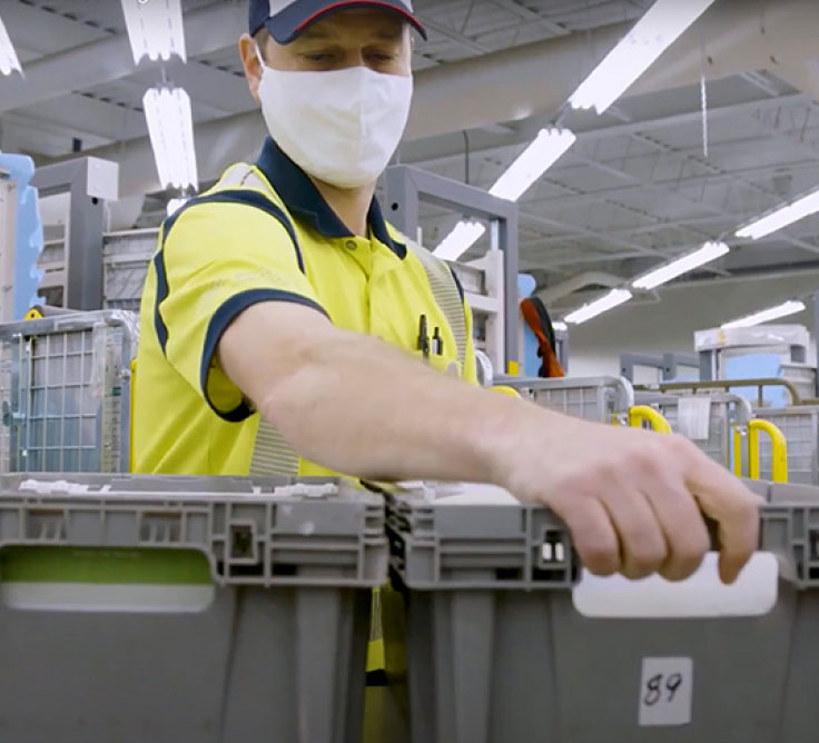 Un employé de Postes Canada porte un gilet de haute visibilité jaune et un couvre-visage. Il s’empare d’un conteneur de courrier dans une installation de tri.