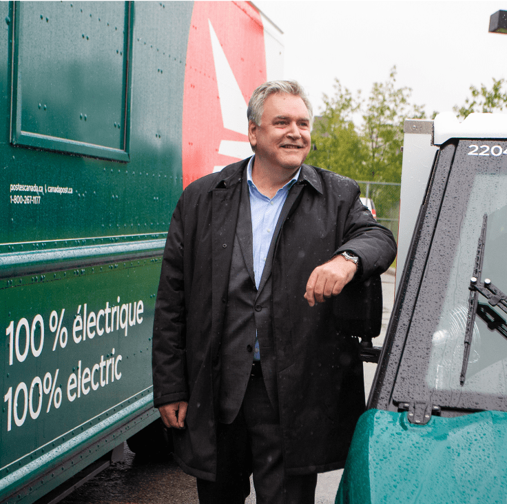Canada Post President and CEO Doug Ettinger stands in between two green fleet vehicles.
