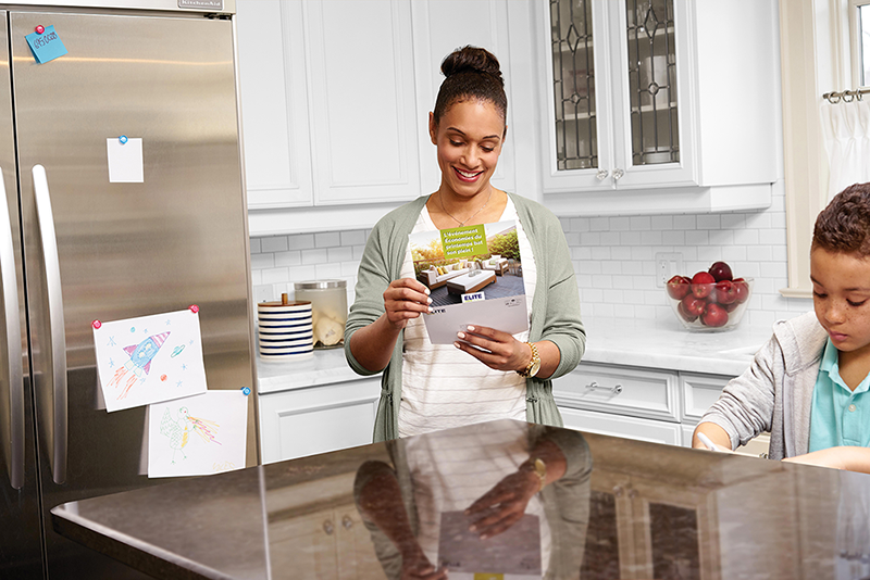 Une femme sourit en lisant une brochure dans sa cuisine. Son enfant se tient &agrave; proximit&eacute;