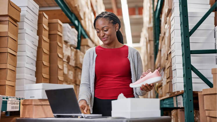 A woman stands at a desk with a laptop and an open shoe box amidst stacks of shoe boxes in a warehouse.