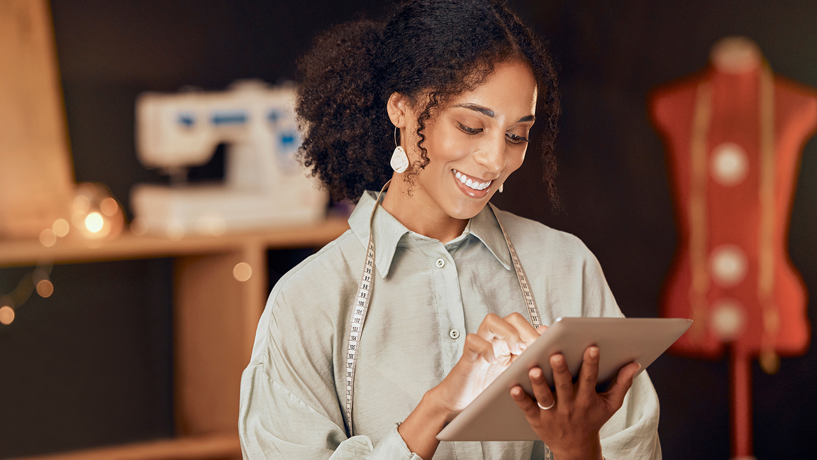 Smiling small business owner in light shirt with measuring tape around neck uses tablet in sewing workspace.