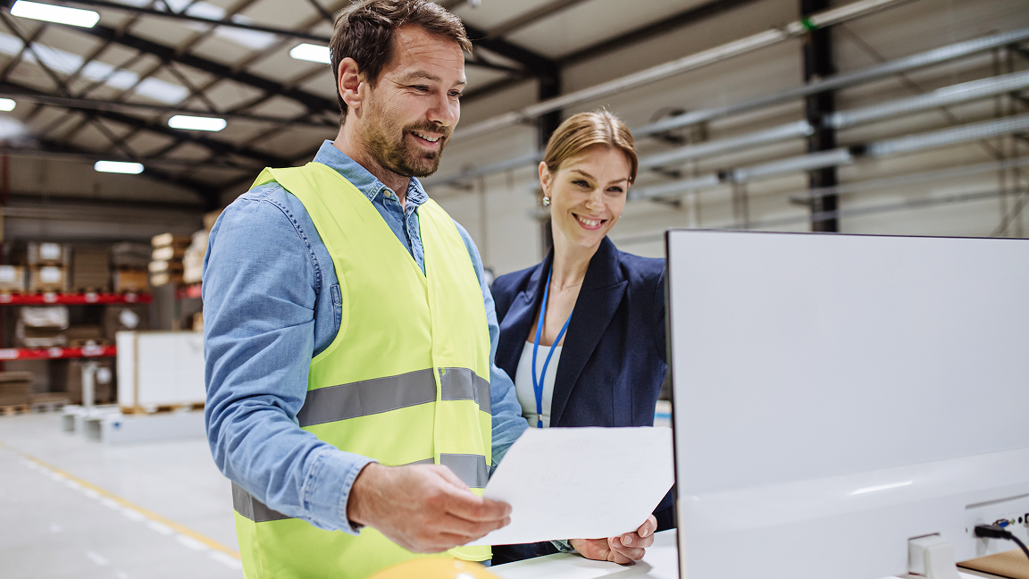 Two people reviewing documents together in an industrial workspace, with one wearing a high‑visibility vest.