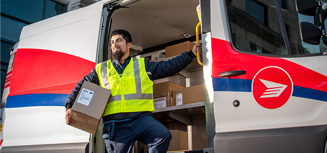 A Canada Post delivery agent in a high-visibility vest carrying a parcel while stepping out of a delivery vehicle loaded with boxes.
