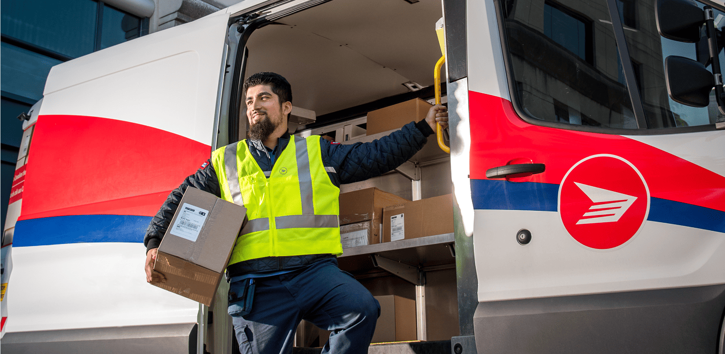 A Canada Post delivery agent in a high-visibility vest carrying a parcel while stepping out of a delivery vehicle loaded with boxes.