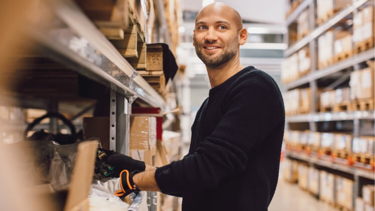 A man smiles as he sorts a box on the shelf of a large warehouse. 