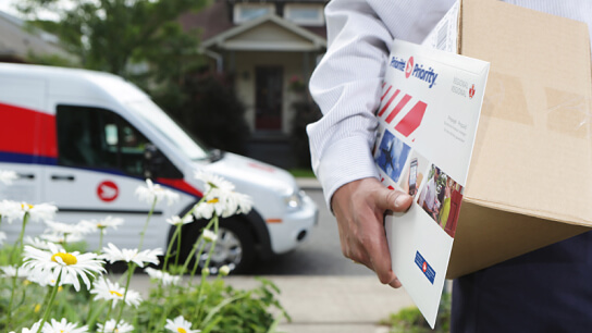A letter carrier holds a shipping box and a bubble mailer. Their Canada Post vehicle is parked nearby. 