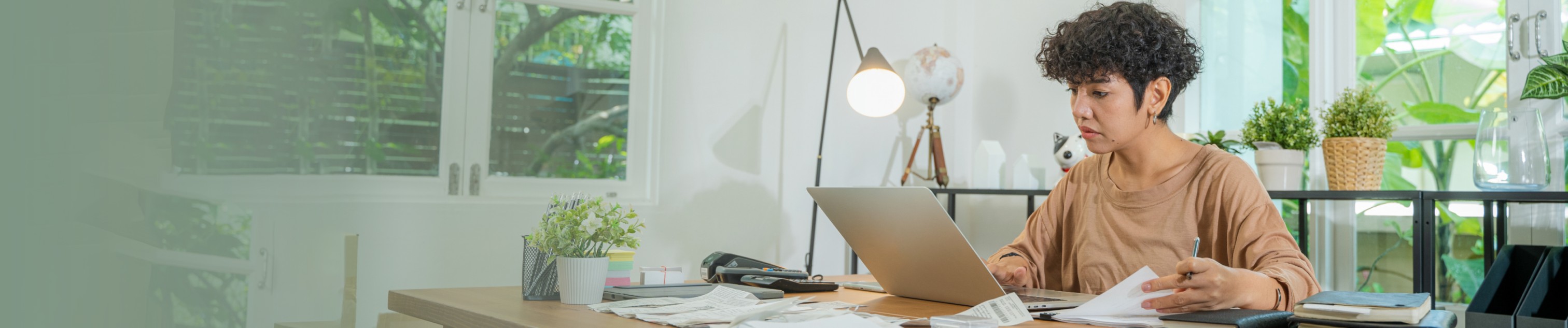 A woman sits at a desk while working on a laptop and flipping through a stack of printed pages. 