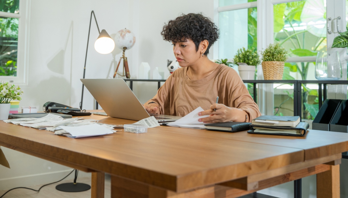 A woman sits at a desk while working on a laptop and flipping through a stack of printed pages. 