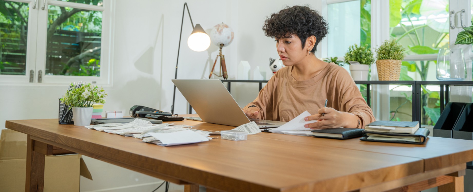 A woman sits at a desk while working on a laptop and flipping through a stack of printed pages. 