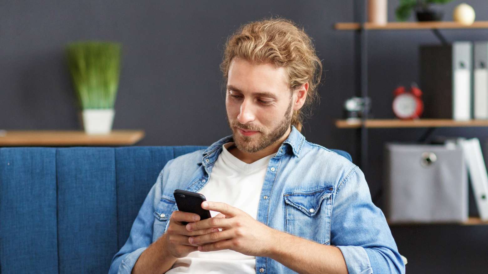 A young man sitting on a couch smiles and holds a mobile phone.