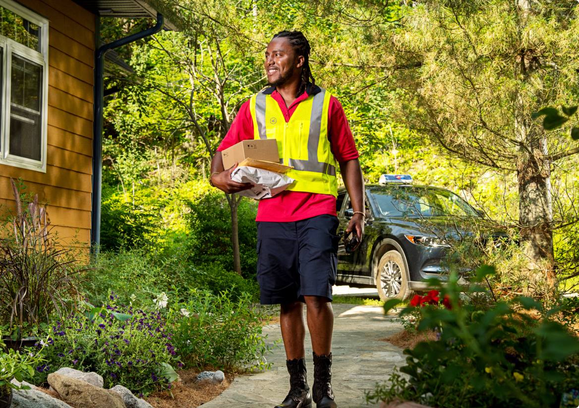 A delivery agent walks along the landscaped path beside a house, carrying packages for delivery.