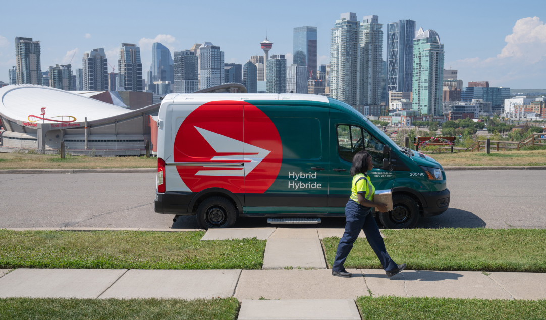 A Canada Post driver walks on a sidewalk and carries a package for delivery. A hybrid Canada Post van is parked behind her on the street. In the background is the Calgary skyline.